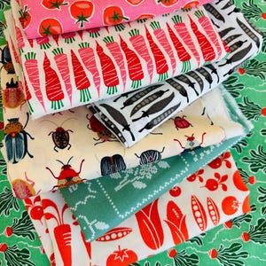 Peut inclure: Une pile de tissus pliés avec des motifs colorés de légumes et d'insectes. Les motifs comprennent des tomates roses, des carottes rouges, des gousses de pois noires et blanches et divers coléoptères. Les tissus sont principalement blancs, roses et verts.