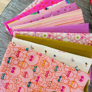 May include: A stack of folded fabric squares featuring various patterns including pink with red stars, purple with strawberries, pink stripes, gold with speckles, white with bees and flowers, and a peach print of stylized faces wearing sunglasses.