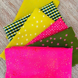 Peut inclure: Un assortiment d'échantillons de tissus colorés aux motifs variés. Les tissus comprennent un rose vif avec des éclats dorés, un jaune avec des étoiles blanches, un motif à carreaux vert et noir et un tissu vert avec des étoiles roses. Les tissus sont disposés sur une surface en bois.