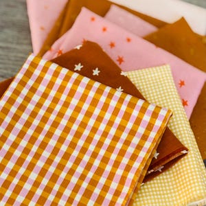 Peut inclure: Un empilement de carrés de tissu aux couleurs variées. Les tissus comprennent du rose, du jaune et du marron, avec des motifs d'étoiles et de carreaux. Idéal pour le patchwork ou d'autres projets créatifs.