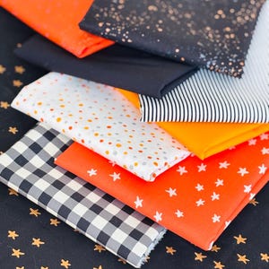 Peut inclure: Un empilement d'échantillons de tissus variés aux tons orange, noir, blanc et gris. Les motifs comprennent des pois, des étoiles, des rayures et un motif vichy. Les tissus sont probablement destinés à des projets de couture.