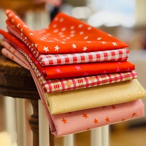 Peut inclure: Une pile de carrés de tissu pliés de différentes couleurs et motifs. Les tissus comprennent du rouge avec des étoiles blanches, du vichy rouge et blanc, du rouge avec des étoiles roses et du rose avec des étoiles orange. Les tissus sont empilés sur une surface en bois.