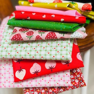 May include: A stack of folded fabric squares featuring various patterns including red strawberries on white, yellow with white stars, solid pink, red with small white strawberries, green with speckles, pink with strawberries, green gingham with tulips, and floral prints.