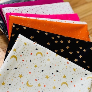 May include: A stack of folded fabric pieces featuring various patterns including gold metallic stars, moons, and speckles on white, black, orange, and magenta backgrounds. The textiles are layered to display the different prints and colors clearly.