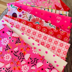 Pode incluir: Uma pilha de tecidos dobrados com vários padrões, incluindo estampas florais em rosa e branco, formas geométricas em vermelho e rosa, estrelas e um tecido rosa brilhante com o texto Little Joys impresso ao lado de pequenas ilustrações de caracóis.