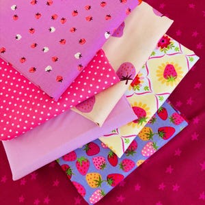 May include: A collection of folded fabric pieces in various shades of pink and purple. Patterns include strawberries, polka dots, and floral designs. The fabrics are arranged on a magenta background with star patterns.