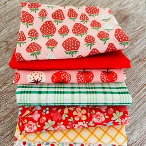 Peut inclure: Une pile de tissus colorés avec différents motifs. Le tissu supérieur présente une impression de fraises sur fond rose. D'autres tissus incluent des motifs rouges, à carreaux, floraux et vichy. Les tissus sont soigneusement pliés et empilés.