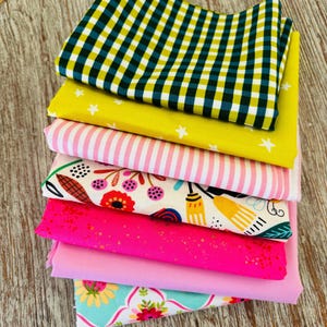 Peut inclure: Une pile de carrés de tissu colorés avec différents motifs. Le tissu supérieur présente un motif à carreaux noirs et jaunes. D'autres tissus incluent du jaune avec des étoiles blanches, des rayures roses, des imprimés floraux et des roses unis. Les tissus sont empilés sur une surface en bois clair.