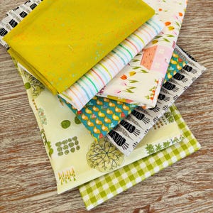 Peut inclure: Une pile de carrés de tissu colorés avec divers motifs. Les tissus comprennent un jaune moutarde avec des mouchetures bleues, du tissu rayé, des imprimés floraux et des motifs géométriques. Les tissus sont disposés sur une surface en bois.