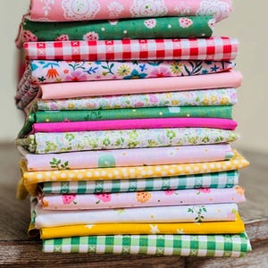 Può includere: Una pila di tessuti colorati con vari motivi. I tessuti includono disegni floreali, a quadri e a pois nei toni del rosa, verde, giallo e bianco. I tessuti sono impilati su una superficie di legno.