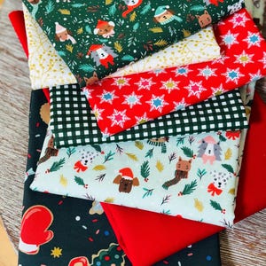 Può includere: Una pila di tessuti piegati con vari motivi natalizi, tra cui cani con cappelli invernali, fiocchi di neve rossi e bianchi, quadretti vichy verdi e bianchi e una stampa verde scuro con camioncini rossi, muffole e omini di pan di zenzero.