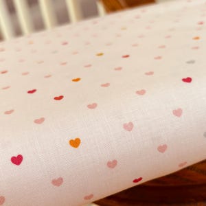 May include: Close-up of a light pink fabric with a repeating pattern of small hearts in shades of red, pink, orange, and gray. The fabric appears to be linen or a similar woven material, with a slightly textured surface.