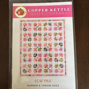 May include: A quilt pattern titled "Leaf Pile" by Copper Kettle Quilt Company. The pattern features a repeating leaf design in shades of pink, orange, green, and gray on a white background. The package includes runner and throw sizes.