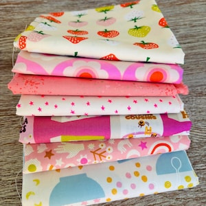 Peut inclure: Une pile de carrés de tissu colorés avec divers motifs. Les motifs comprennent des fraises, des fleurs, des étoiles et le texte "COUSINS". Les tissus sont dans des tons de rose, blanc et bleu.