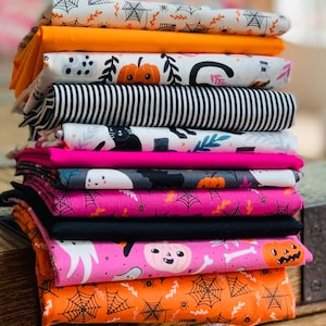 Puede incluir: Una pila de 10 cuadrados de tela con temática de Halloween con varios patrones, incluyendo telarañas naranjas y negras, fantasmas rosas, gatos negros y calabazas. Las telas están dispuestas en una exhibición colorida y festiva.