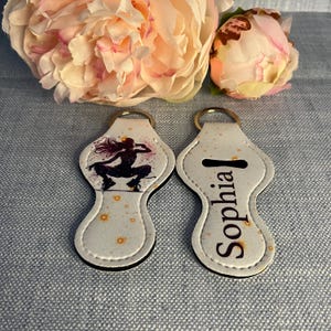 May include: Two white lip balm holders with keyrings. One holder features a silhouette of a woman dancing against a white and orange background. The other holder has the name "Sophia" written in black.