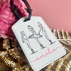 Peut inclure: Étiquette de bagage blanche avec un dessin au trait noir de trois danseuses de ballet. L'étiquette a une sangle marron et le nom "amelia" en script rose. Le fond est rose et doré.