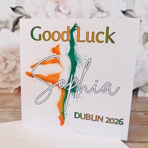 May include: White greeting card with the words "Good Luck Sophia" and "Dublin 2026". The card features a dancer in orange and green, with a green and orange color scheme. The card is on a wooden surface.