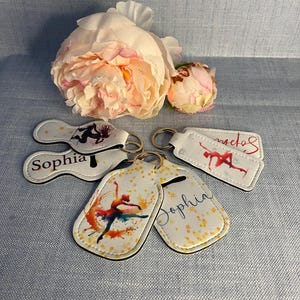 May include: Four white keychains with different designs. One keychain features a ballerina with a watercolour background, another has a silhouette of a dancer, and two have the name "Sophia" written on them. The keychain with the ballerina has gold glitter accents.