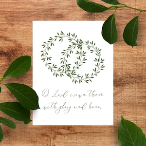 May include: A white print with two green leaf wreaths intertwined, with the text "O Lord crown them with glory and honor" in a script font. The print is on a wooden surface with green leaves.
