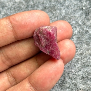 Peut inclure: Une pierre précieuse de rubis brute et non taillée, d'une teinte rouge profond. La pierre naturelle présente une surface texturée et une forme irrégulière, mettant en valeur son état brut et non poli. Le rubis est tenu dans une main.
