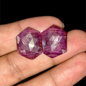 Pode incluir: Duas pedras preciosas de rubi facetadas, cada uma com forma hexagonal e uma cor rosa intensa. As pedras têm um brilho ligeiramente metálico e estão colocadas sobre um fundo neutro.