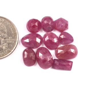 May include: A selection of faceted, deep red ruby gemstones in various shapes, including oval, teardrop, and rectangular cuts. A US quarter coin is included for size comparison, set against a white background.