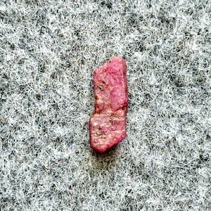 May include: A rough-cut, natural ruby gemstone. The ruby is a vibrant pinkish-red color with a slightly irregular shape. The gemstone has a textured surface and appears to be unpolished, showcasing its raw, natural beauty. The background is a textured gray.