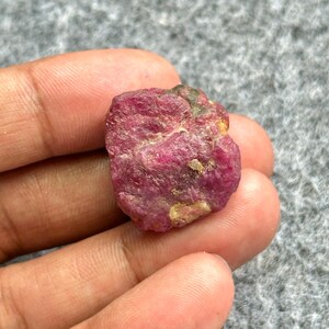 May include: A rough, uncut ruby gemstone with a deep red hue. The surface has a textured appearance with some lighter-colored areas. The gemstone is held in a hand against a gray background. This natural ruby is a raw mineral specimen.