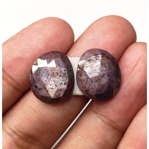 Pode incluir: Duas pedras preciosas ovais facetadas de cor roxa e cinza. As pedras preciosas têm um aspeto salpicado.