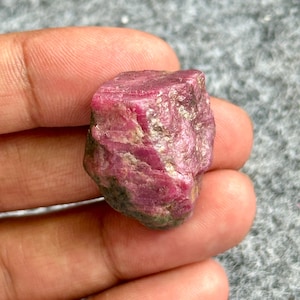Könnte beinhalten: Ein roher, ungeschliffener Rubin-Edelstein mit tiefroter Farbe. Der Naturstein hat eine strukturierte Oberfläche mit sichtbaren kristallinen Strukturen und einigen helleren Bereichen. Der Rubin wird in einer Hand gehalten, um seine Größe und Form zu zeigen.
