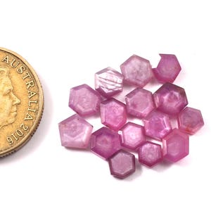 Pode incluir: Uma coleção de 16 gemas hexagonais facetadas cor-de-rosa, cada uma com o tamanho aproximado de uma moeda de 10 cêntimos de euro. As gemas estão dispostas em um monte solto sobre uma superfície branca.