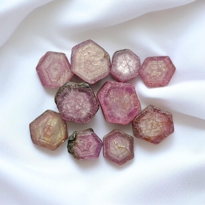 Puede incluir: Una colección de piedras preciosas facetadas de color rosa y verde, con forma hexagonal, expuestas sobre un fondo de tela blanca.