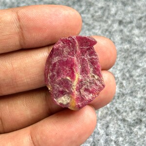 May include: A raw, uncut ruby gemstone in a deep red colour. The natural stone has a textured surface with lighter, yellowish-white inclusions. The ruby is held in a hand, showcasing its raw, organic form.