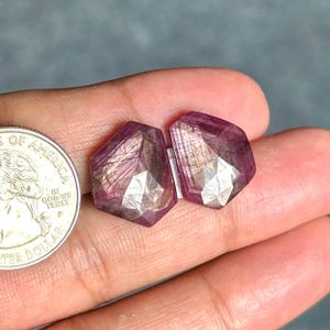 May include: Two faceted, shield-shaped, deep magenta-colored gemstones with visible internal inclusions and a polished surface, positioned next to a United States quarter coin for scale to show their relative size.