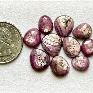 May include: A collection of faceted, oval-shaped, deep pink gemstones with internal patterns, next to a U.S. quarter coin. The gemstones vary in size and shape, with a polished, reflective surface.