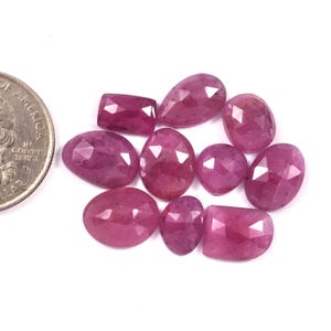 May include: A selection of faceted ruby gemstones in various shapes, including oval and rectangular. The gemstones are a deep pinkish-red hue and are presented on a white background. A coin is included for size comparison.