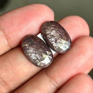 Pode incluir: Duas pedras preciosas ovais facetadas com um tom roxo escuro e manchas prateadas. As pedras estão colocadas sobre um fundo rosa claro.