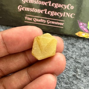 May include: A small, yellow gemstone with a geometric shape is held between fingers. The gemstone has a textured surface and appears to be a natural stone. The background includes text that reads "Gemstone Legacy Co" and "Fine Quality Gemstone."