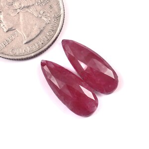 May include: Two faceted, teardrop-shaped, deep red gemstones are displayed next to a quarter. The gemstones have a polished, reflective surface and are set against a white background. The gemstones are approximately 1 inch in length.