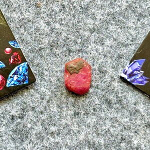 May include: A close-up of a red ruby gemstone with a dark gray inclusion. The ruby is rough and unpolished, with a natural, irregular shape.