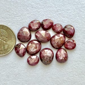 May include: A collection of faceted, oval-shaped, and irregularly shaped ruby gemstones. The gems are a deep red color with reflective surfaces. A US quarter is included for size comparison. The gemstones are arranged on a white surface.