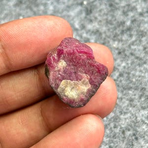 May include: A rough, uncut ruby gemstone. The ruby is a deep, vibrant red color with lighter, off-white patches. The stone has a raw, natural appearance, with an uneven surface and a dark, almost black, border.