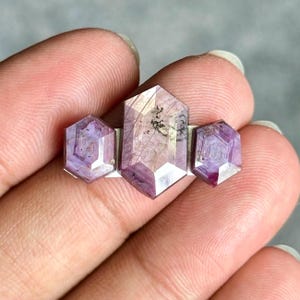 May include: A ring featuring three hexagonal-cut purple gemstones set in a silver-toned metal band. The central stone is larger than the two flanking stones, and all three display natural internal inclusions and faceted surfaces.
