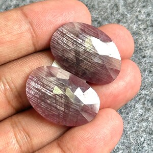 Puede incluir: Dos gemas ovaladas y facetadas con un tono rosa púrpura. Las piedras tienen un patrón texturizado y lineal y se sostienen en una mano sobre un fondo gris. Las gemas son probablemente para la fabricación de joyas.