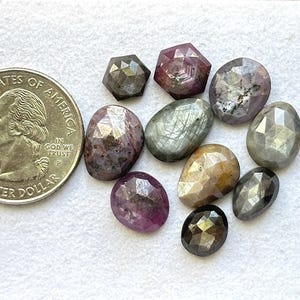 Op de afbeelding: Een verzameling gefacetteerde edelstenen in verschillende vormen en kleuren, waaronder roze, grijs en bruin. Een Amerikaanse kwartmunt is inbegrepen ter vergelijking van de grootte. De edelstenen worden op een wit oppervlak weergegeven.