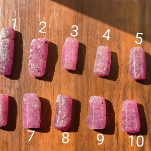 Op de afbeelding: Tien roze robijnstenen, genummerd van 1 tot en met 10, gerangschikt in twee rijen op een houten oppervlak. De stenen zijn allemaal rechthoekig van vorm en hebben een ruwe, natuurlijke textuur.