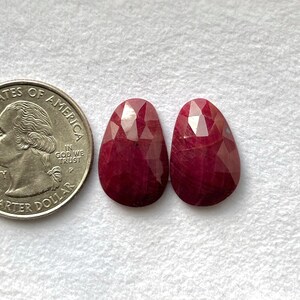May include: Two faceted, oval-shaped, deep red gemstones are displayed next to a U.S. quarter. The gemstones have a polished, reflective surface. The quarter provides a size reference.