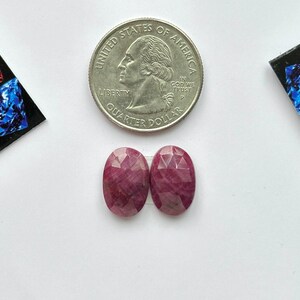 May include: Two oval-shaped, faceted pinkish-red rubies positioned below a United States quarter coin for scale. The gemstones exhibit a translucent quality with natural internal inclusions and a polished surface, reflecting light across their cut facets.
