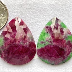 May include: Two faceted, teardrop-shaped gemstones. The gems feature a vibrant mix of deep red and green hues, with a quarter coin for size comparison. The gemstones are set against a white background.